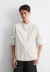 Chemise blanche à boutons, avec des pois noirs, col mandarin, manches longues retroussées aux poignets, et coupe droite. Portée avec un pantalon large foncé.