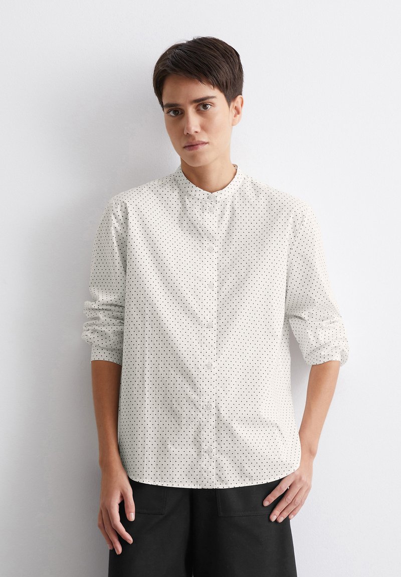Chemise blanche à boutons, avec des pois noirs, col mandarin, manches longues retroussées aux poignets, et coupe droite. Portée avec un pantalon large foncé.