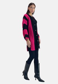 Gehaakte cardigan in roze en zwarte strepen, met een knopenfront, lange mouwen en een gestructureerde afwerking. Gecombineerd met zwarte strakke broek en laarzen tot de knie.