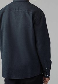 Chemise à manches longues en tissu sombre tissé avec un motif texturé, poignets boutonnés et col de chemise. Le tissu semble respirant et léger.