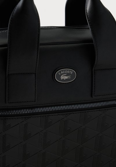 Sac Lacoste noir avec motif géométrique texturé, deux poignées, fermeture éclair et logo ovale avec design de crocodile.