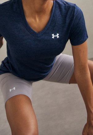 Persoon draagt een marineblauw V-hals T-shirt en lichtgrijze shorts met Under Armour-logo's, in een lage hurk- of stretchpositie.