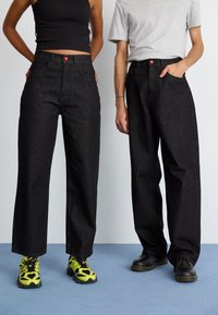 Sorte denim vide bukser med høj talje, med røde knap detaljer. Båret med kontrasterende fodtøj: lysende grønne sneakers og sorte sko.