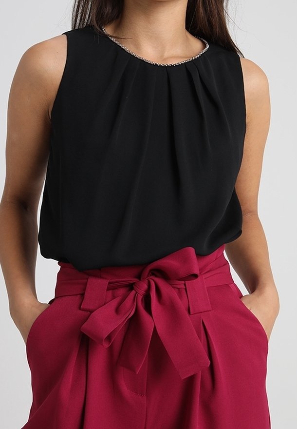 Svart ärmlös topp med plisserad design och pärlbesatt halsringning, matchad med högmidjade vinröda byxor med knytband i fronten.