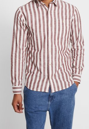 Mand iført brun og hvid lodret stribet skjorte med brystlomme og blå jeans, den ene hånd i lommen, på ensfarvet baggrund.