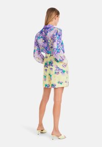 Blumenbluse in Lila und Blau mit langen Ärmeln, kombiniert mit einem gelben Rock mit lila Blumenmustern. Die Accessoires umfassen blassgelbe Heels.