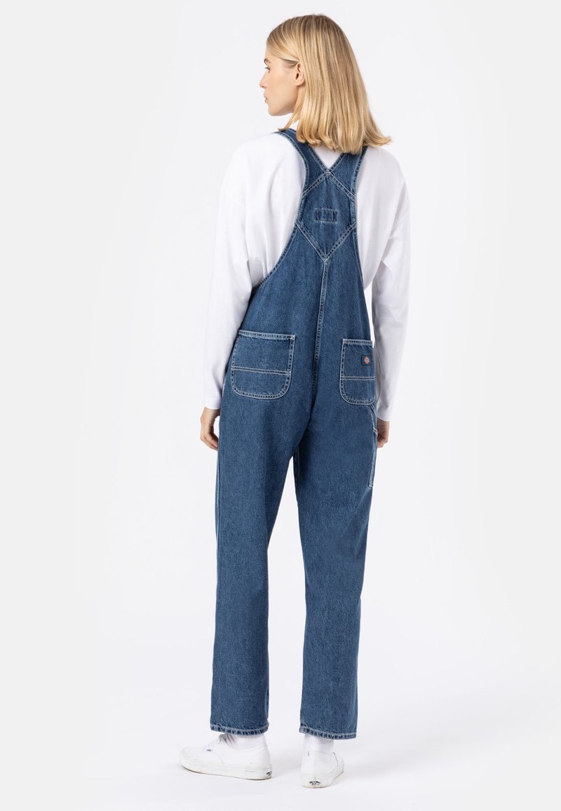 Salopette en denim bleu avec bretelles réglables, dotée de deux poches arrière et d'une coupe décontractée, associée à un haut blanc à manches longues et des baskets.