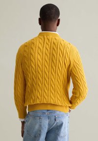 Gelber Kabelstrickpullover mit geripptem Kragen und Saum. Mit strukturierten Mustern und cropped Design. Getragen über hellblauen Jeans.