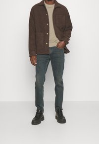 Chaqueta marrón texturizada con dos bolsillos frontales sobre un suéter de punto beige, combinada con vaqueros azules y botas negras con cordones.