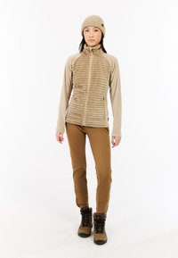 Beige fleecedonsjack met verticale ribbeling, gecombineerd met tan geklede broek en bruine wandelschoenen. Een gebreide beige beanie wordt op het hoofd gedragen.