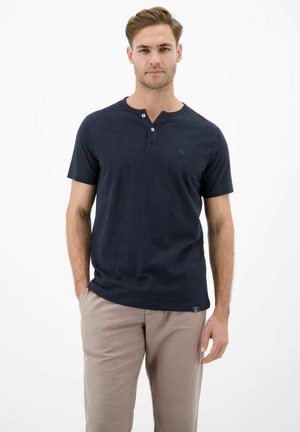 Mann mit hellbraunem Haar, der ein marineblaues Henley-Shirt mit kurzen Ärmeln und hellbeige Hosen trägt, vor einem schlichten weißen Hintergrund stehend.