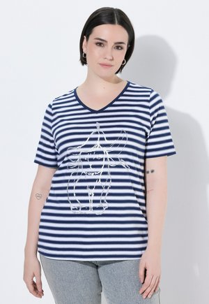 Kobieta z krótkimi ciemnymi włosami, ubrana w granatowo-biały t-shirt w paski z dekoltem w serek i srebrnym abstrakcyjnym kwiatowym wzorem, stoi na białym tle.