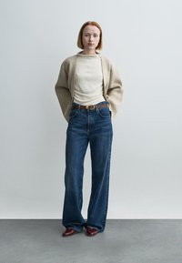 Μπεζ ζακέτα πάνω από ένα κρεμ μπλουζάκι με μακριά μανίκια, σε συνδυασμό με τζιν παντελόνι ψηλόμεσο σε μπλε απόχρωση. Καφέ ζώνη και καφέ παπούτσια. Μονόχρωμα ρούχα με άνετη εφαρμογή.