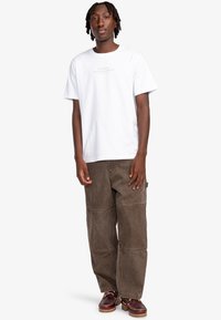 Camiseta blanca de algodón con "ELEMENT SKATEBOARD CO." impreso en gris. Pantalones de pana marrón con bolsillos en la parte delantera y un ajuste relajado. Zapatos marrones.