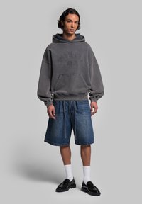 Sweat à capuche gris surdimensionné avec poche kangourou, présentant des lettres en relief. Associé à un short en denim à longueur de genou et des mocassins noirs.