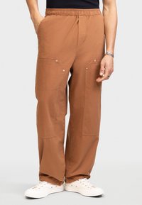 Brown cargo pants with an elastic waistband, featuring two large pockets on the thighs, snap buttons, and a relaxed fit, paired with white sneakers.