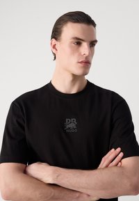 T-shirt in cotone nero con maniche corte, caratterizzato da un logo goffrato discreto sul lato sinistro del petto. Tessuto liscio, scollatura rotonda.