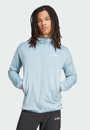 Mand med lange dreadlocks iført lyseblå Terrex lynlåstrøje og sorte Terrex bukser, stående med hænderne i lommerne.