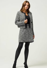 Cappotto cropped grigio e nero con texture abbinato a una gonna. Presenta dettagli neri a tinta unita. Indossato con collant neri e tacchi.