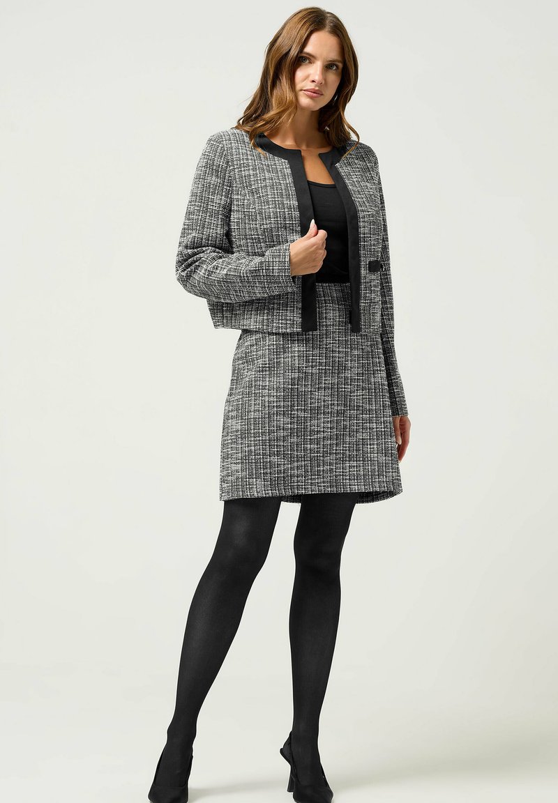 Cappotto cropped grigio e nero con texture abbinato a una gonna. Presenta dettagli neri a tinta unita. Indossato con collant neri e tacchi.
