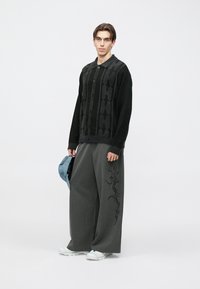 Czarny sweter w wzory z guzikami, szare spodnie o szerokich nogawkach w delikatne prążki z boczną haftowaną ozdobą oraz jasnoniebieska czapka w ręku.