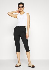 Top de tanque branco cortado com bainha em forma de ondas e três botões, combinado com calças pretas cortadas e sandálias bejes sem fecho.
