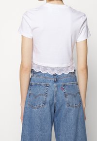 Vitt croppat t-shirt med spetsdetalj. Kombinerat med högmidjade blå denimjeans. Enkla, avslappnade design med texturerad spets.