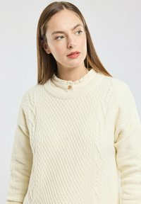 Pull en tricot crème clair avec un motif en cable texturé, col arrondi et un détail de bouton au cou. Tissu épais, coupe décontractée.