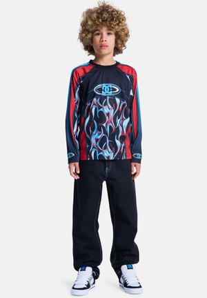 Jongen met krullend haar, gekleed in een langarmig shirt met vlammenpatroon, zwarte broek en witte sneakers, staande tegen een effen achtergrond.