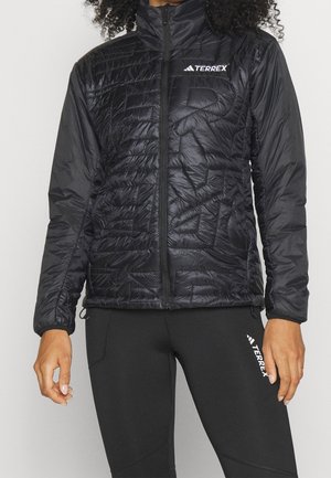 Personne portant une veste matelassée noire Terrex avec une fermeture éclair frontale et des leggings noirs Terrex, debout devant un fond uni.