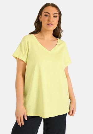 Kvinde iført en lysegul kortærmet V-hals top med sorte bukser, stående mod en ensfarvet hvid baggrund.