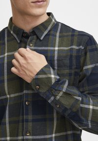 Hombre con camisa de cuadros en verde, azul y blanco abrochándose el primer botón cerca del cuello.