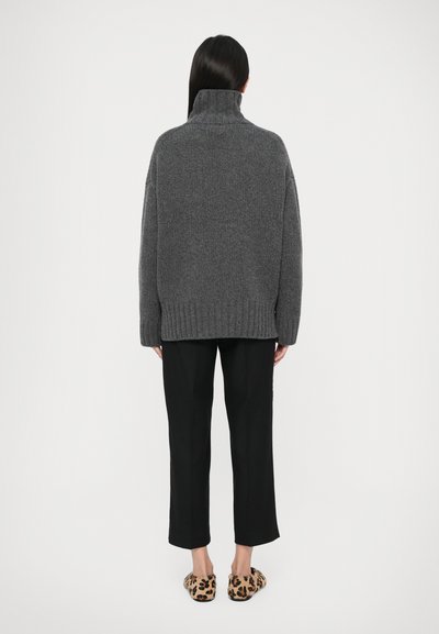 Persona con capelli lunghi e scuri che indossa un maglione grigio a collo alto, pantaloni cropped neri e pantofole maculate, in piedi di spalle.
