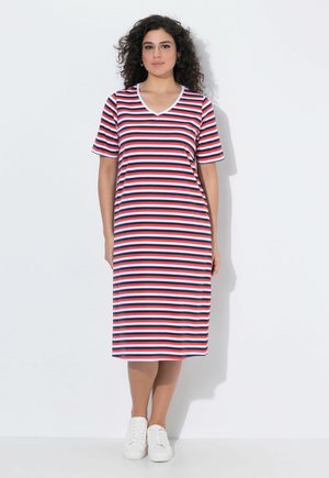 Kvinde stående iført en knælang, kortærmet kjole med vandrette røde, hvide og blå striber og hvide sneakers, mod en hvid baggrund.