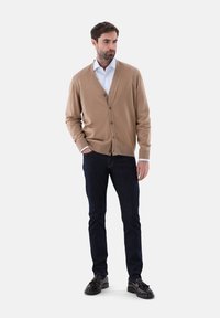 Beige Strickjacke aus einem gestrickten Stoff, mit V-Ausschnitt, Knopfleiste und gerippten Bündchen, kombiniert mit dunklen Jeans und schwarzen Schuhen.
