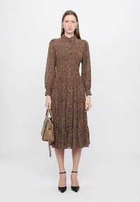 Abito marrone a pois con maniche lunghe, chiusura frontale a bottoni e gonna a più strati. Abbinato a una borsa beige e tacchi neri.