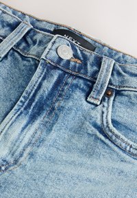 Světlé modré džíny s jemně vybledlým vzhledem, stříbrným knoflíkovým zapínáním, pevnými švy a detaily kapes viditelnými na pasu.