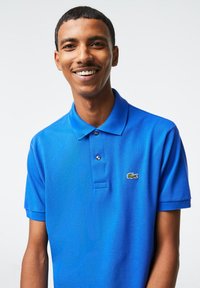 Blaues Poloshirt aus strukturiertem Stoff, mit einer Drei-Knopf-Leiste und umgeschlagenen Ärmeln. Mit grünem Lacoste-Logo auf der linken Brust.