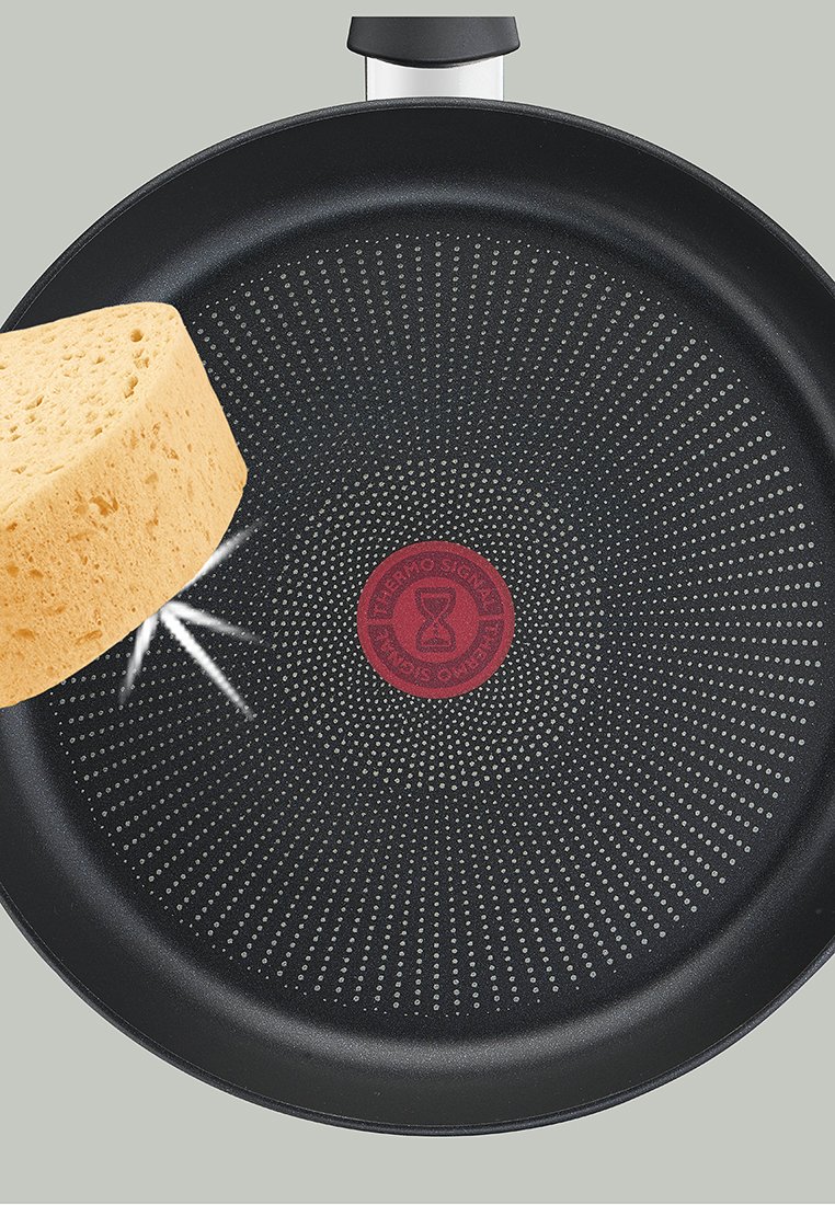 Schwarze antihaftbeschichtete Pfanne mit einem roten kreisförmigen Temperaturindikator in der Mitte, mit einem gepunkteten Muster und einem silbernen Griff. Ein Stück Schwammbrot ruht am Rand.