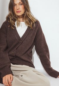 Cardigan marrone lavorato a maglia con scollo a V, dotato di grandi bottoni, maniche larghe e una finitura testurizzata, abbinato a una maglietta bianca e pantaloni crema.