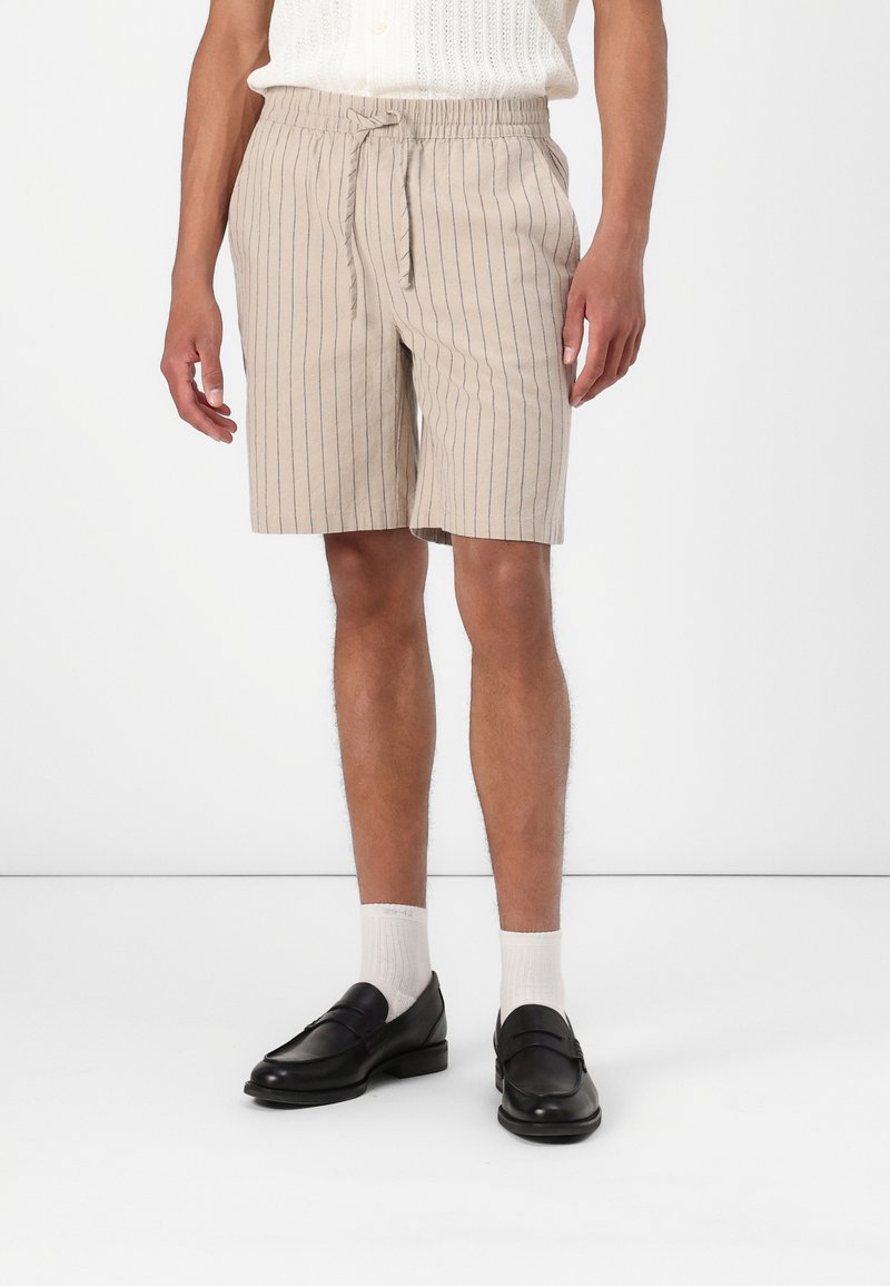 Lys beige shorts med lodrette navy striber, elastisk taljebånd med snøre, parret med sorte loafers og hvide ankelsokker.