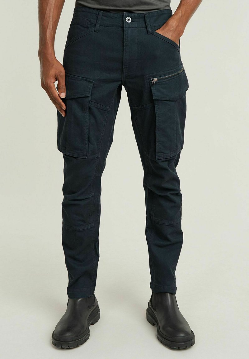 Ciemnoniebieskie spodnie cargo z wieloma kieszeniami, zapinane na zamek i z klapami, wykonane z wytrzymałego materiału, charakteryzujące się zwężającymi się nogawkami i zapięciem na guzik.