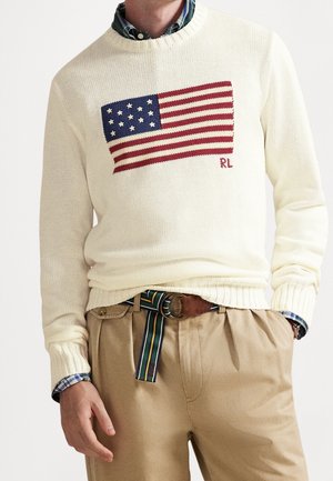 Homme portant un pull crème avec un motif de drapeau américain, une chemise à carreaux en dessous, un pantalon beige et une ceinture en tissu rayé avec un détail en cuir.