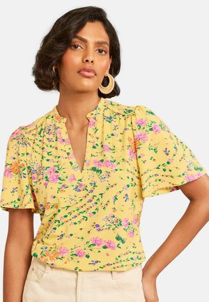Woman wearing yellow short-sleeve blouse with pink and orange floral pattern, ruffled neckline, and beige pants, standing with one hand on hip.