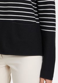 Schwarz-weißer gestreifter Pullover mit strukturierter Oberfläche, ausgestattet mit einer Fronttasche und langen Ärmeln, kombiniert mit hellen Hosen.