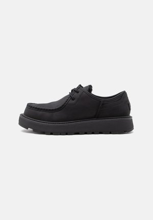Zapato negro de piel con cordones, suela gruesa de goma y detalle cosido en la punta, mostrado de lado sobre un fondo blanco.