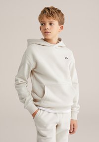 Vaaleanbeige huppari, jossa on kengurutasku, ribbaatut hihansuut ja pieni musta logo rinnassa, yhdistettynä matching-housuihin. Pehmeä kangas.