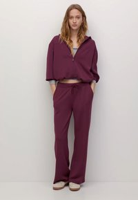 Bluza w kolorze jagodowym z suwakiem z przodu i kapturem, zestawiona z dopasowanymi spodniami o szerokich nogawkach. Oba elementy mają miękkie, teksturowane wykończenie.