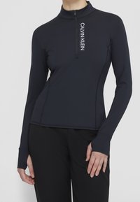 Žena ve fitted černém dlouhém rukávu topu Calvin Klein se čtvrtzipem a otvory pro palce, v kombinaci s černými kalhotami.