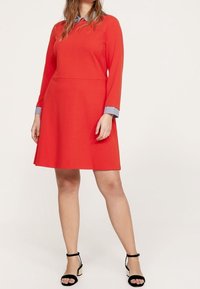 Rotes langärmeliges Kleid mit tailliertem Oberteil, ausgestelltem Rock und einem farblich abgesetzten blauen gestreiften Kragen. Kombiniert mit schwarzen Sandalen mit Knöchellänge.